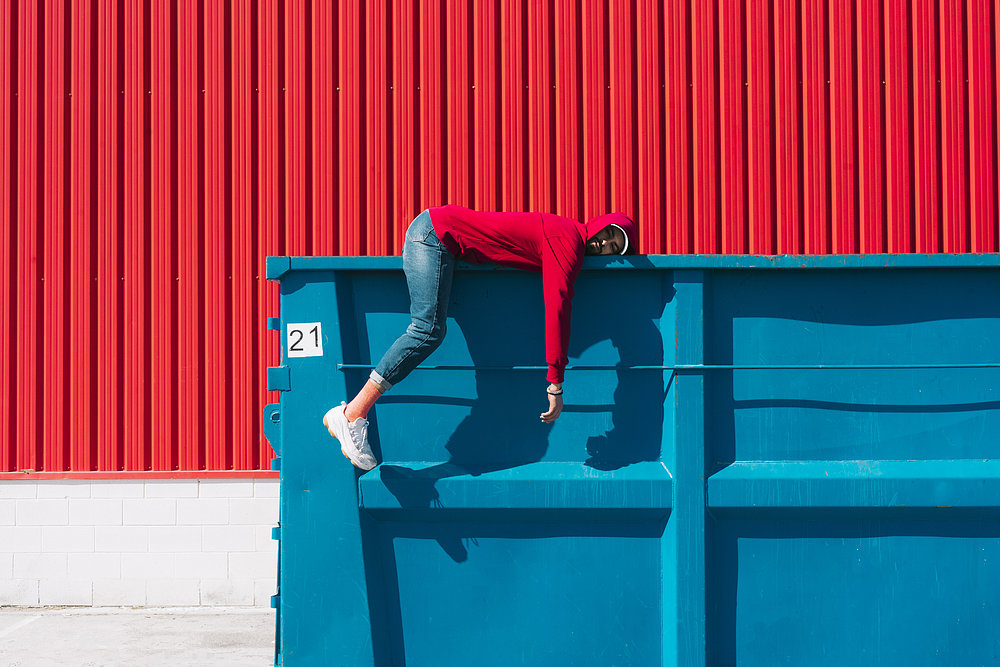 Mann liegt auf einem blauen Container vor einer roten Wand aus lauter Langeweile.